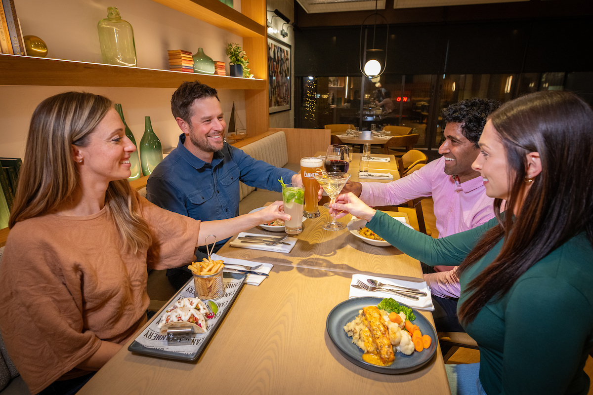 Four people are sitting at a table, dining out at a restaurant. The group is holding their glasses to cheers.