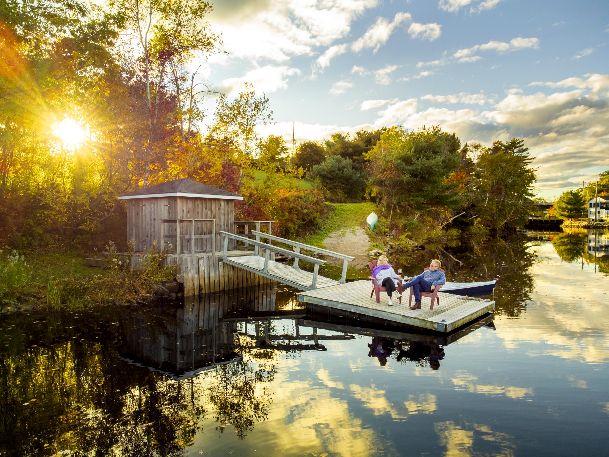 Two people sit in red Adirondack chairs at the end of a wharf. They both hold wine glasses. The sun is setting and the light is casting a perfect reflection of the sky in the water. Everything is lush and green.