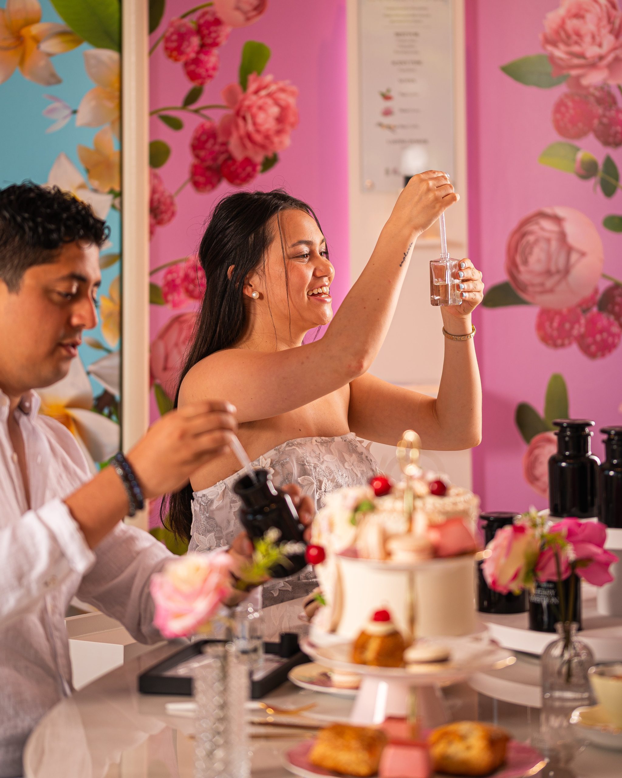 Two people sit in a room with multi coloured walls (pink, blue and flowers). The two people are using droppers to take tiny amounts of liquid from a bottle.