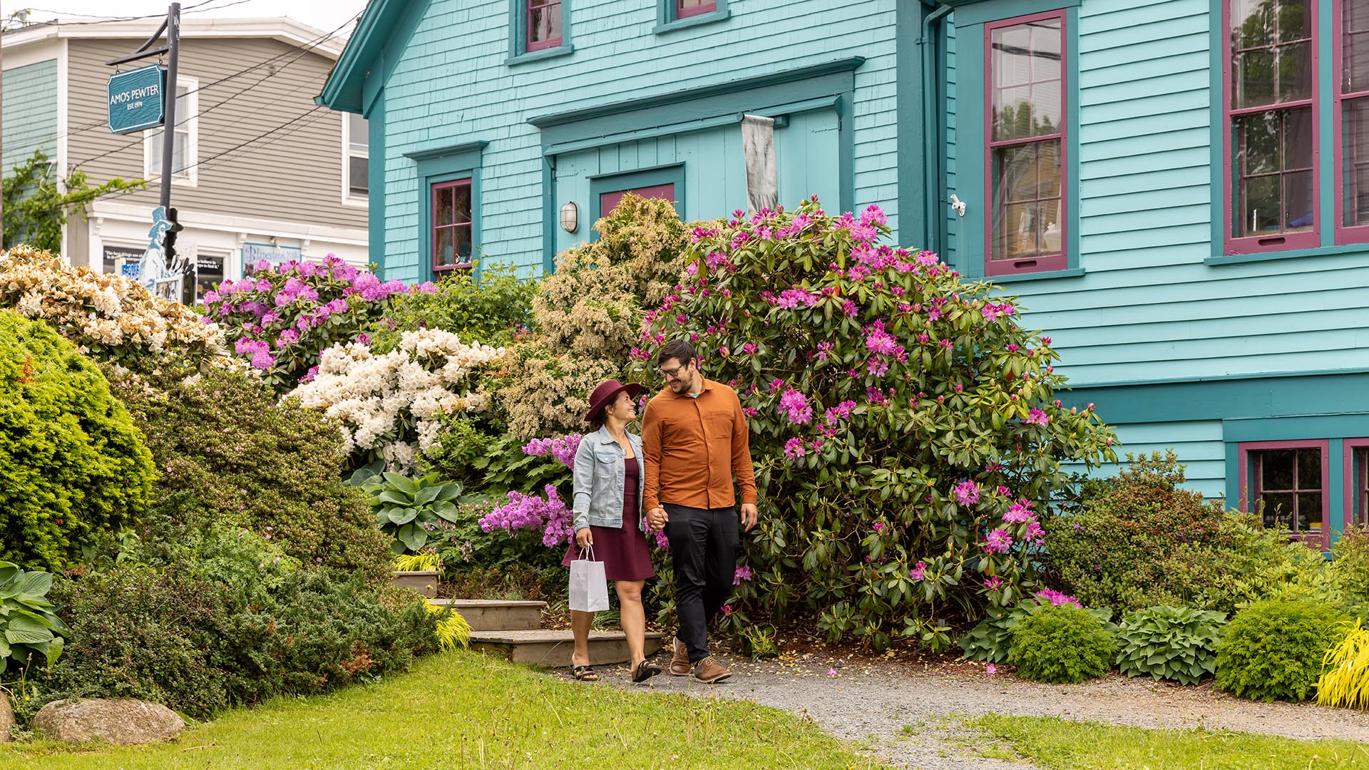 Two people holding hands and smiling at each other outside of Amos Pewter in Mahone Bay Nova Scotia in the summertime with the colourful garden flowers in bloom
