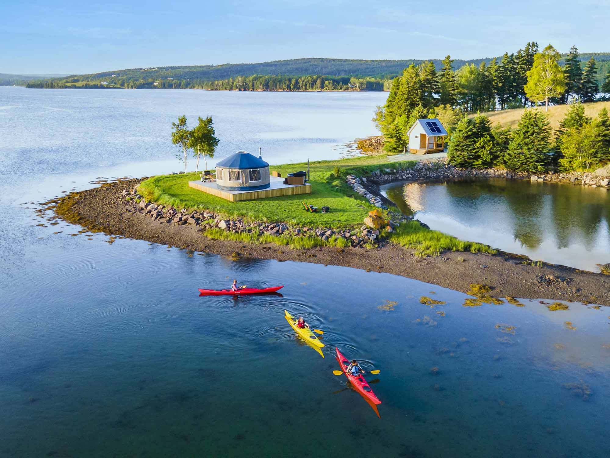 Three people kayaking in colourful sea kayaks on Chedabucto Bay alongside a yurt at Authentic Seacoast Yurts in the Vineyard in the town of Guysborough on Nova Scotia's Eastern Shore.