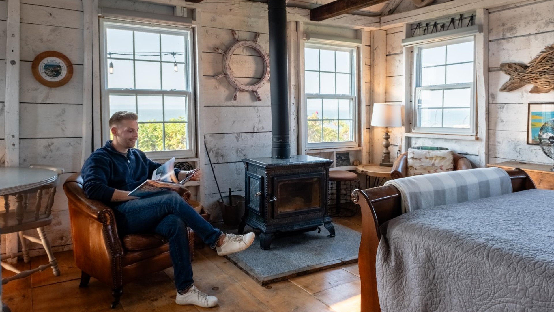 A man sits in a chair looking at a book with scenic ocean views out the window behind him.