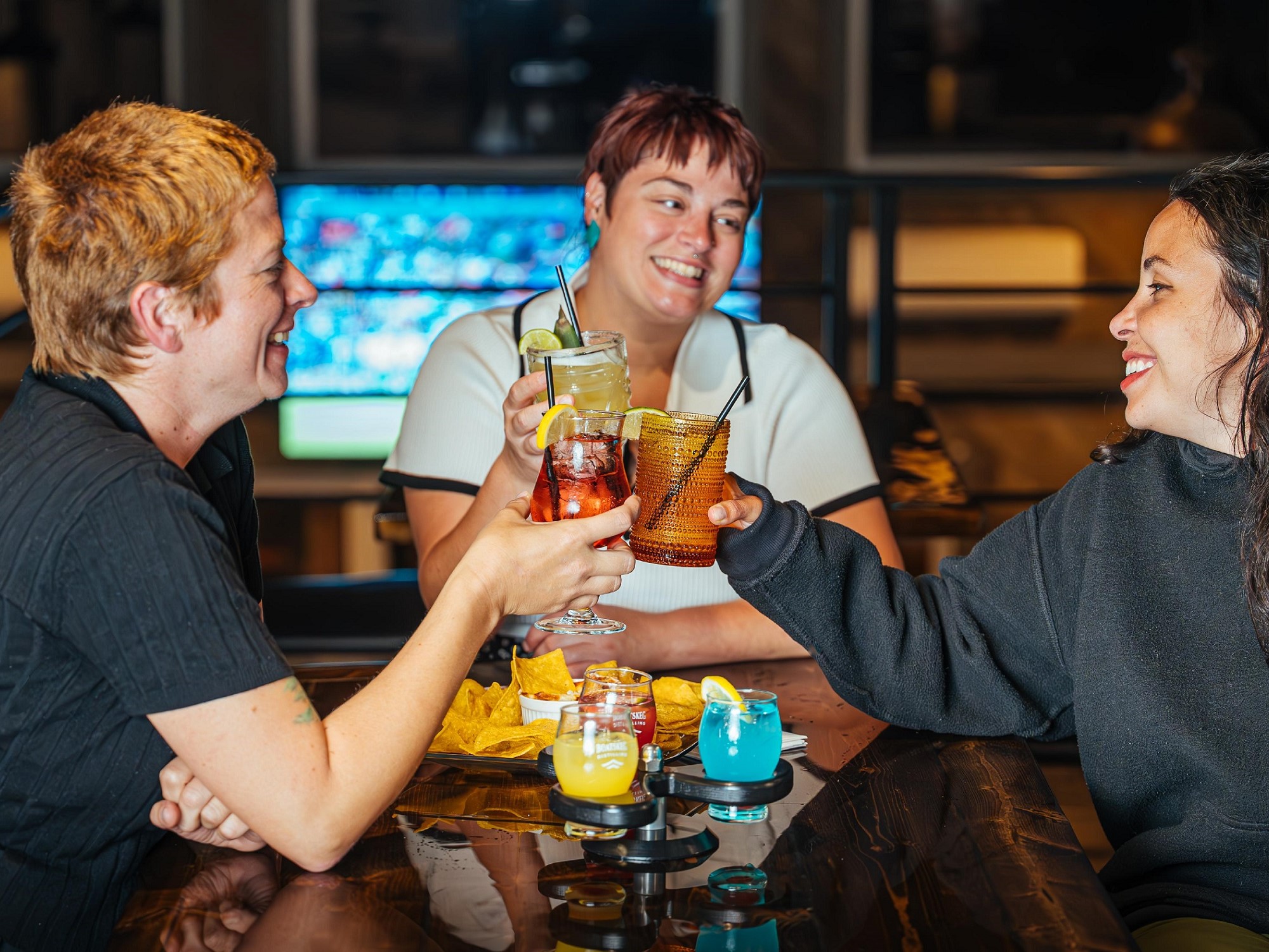 Three friends do a cheers with their glasses of cocktails in a distillery.