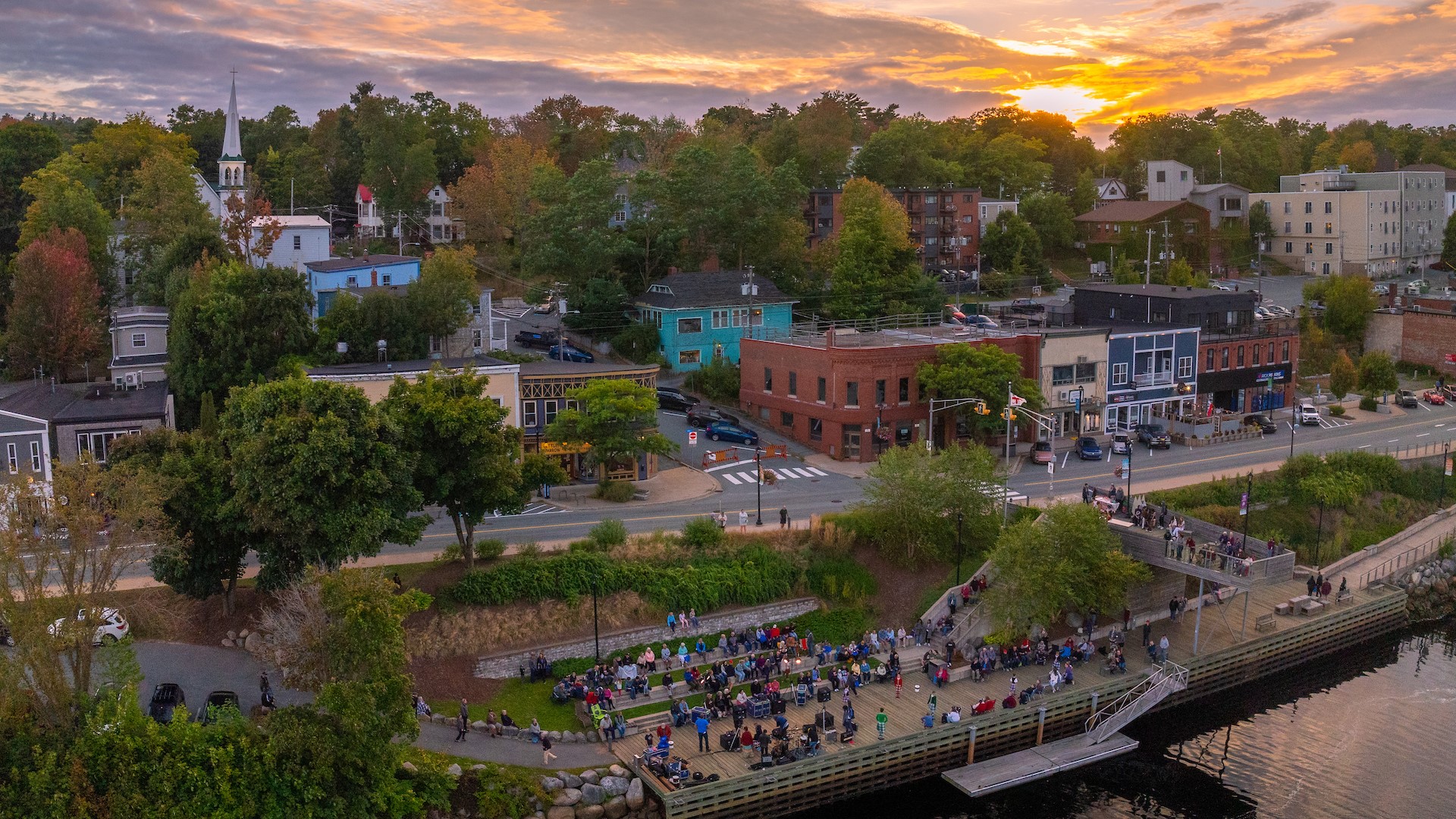 An aerial view of a town at sunset with people gathered along a river boardwalk listening to a live band play.