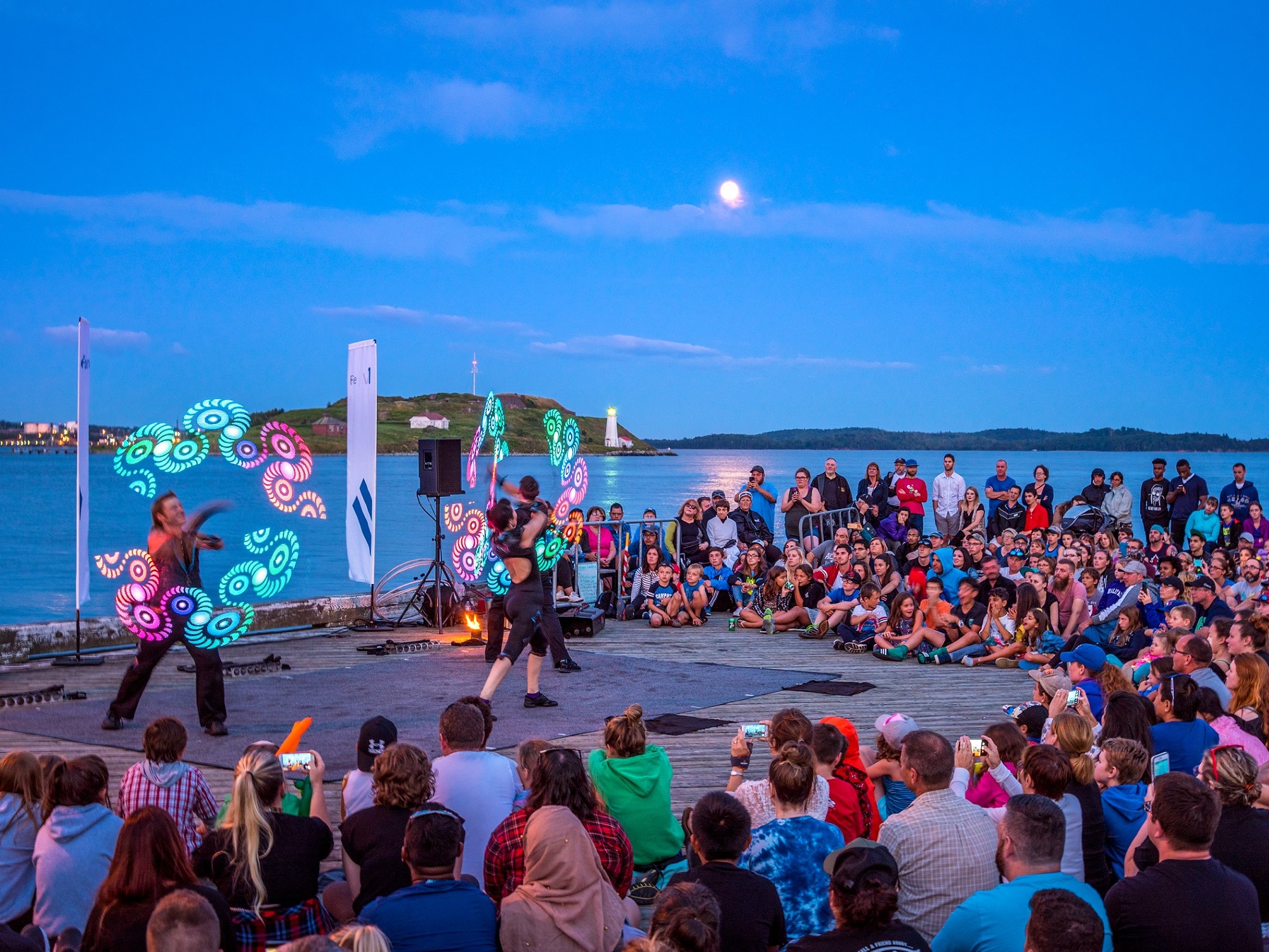 A crowd watches as buskers perform on the boardwalk by the water during evening time.