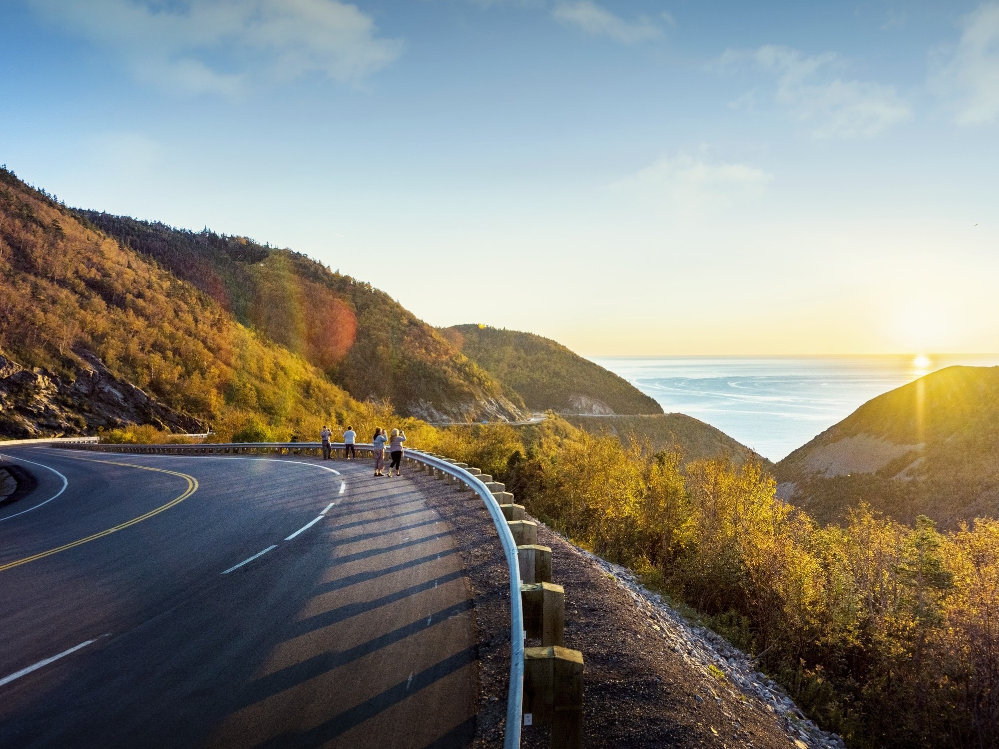 People stand on the side of the road at a scenic view point looking at the sunset