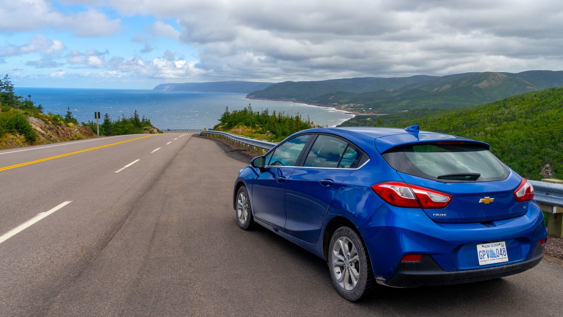 A blue car is parked at a look off with a view of the water and Cape Breton Highlands
