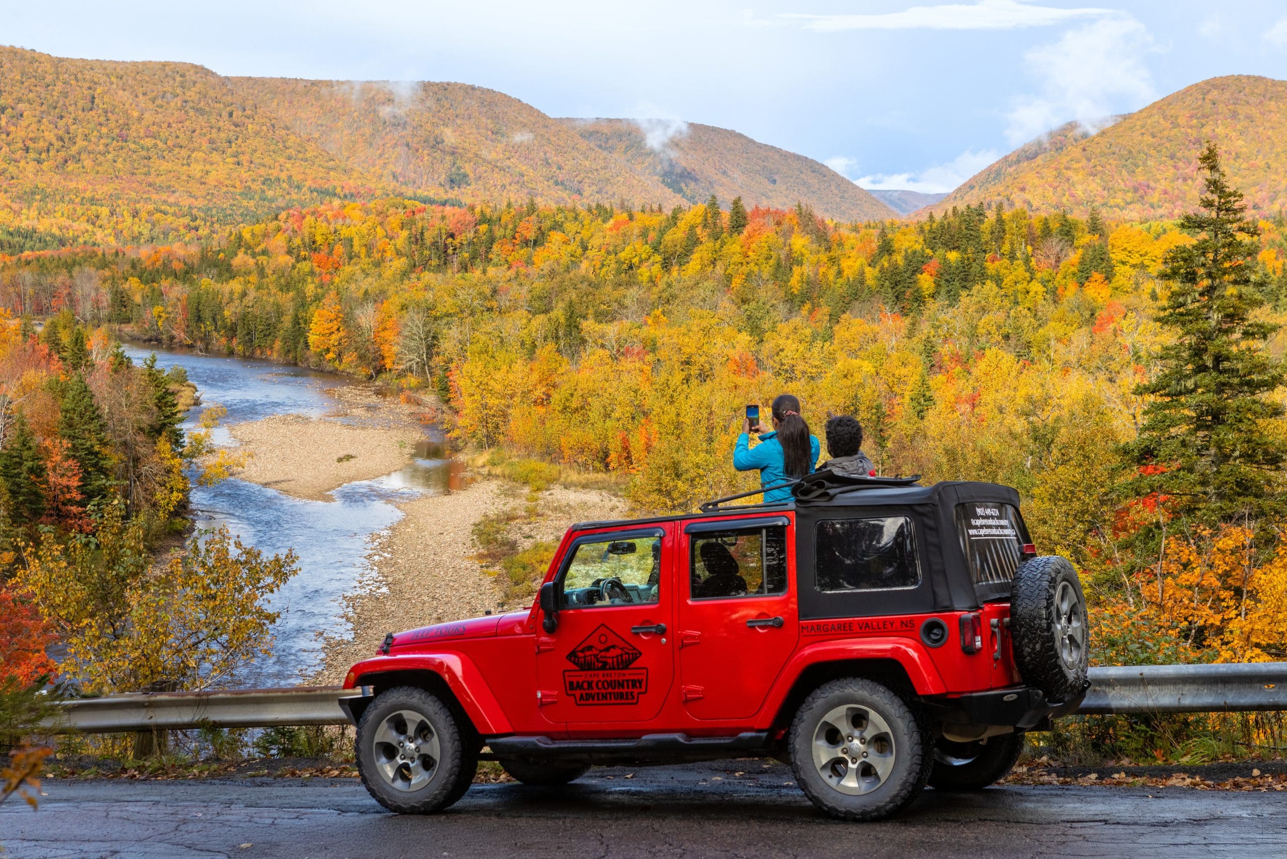 A bright red keep is stopped on the side of the road with two people standing through the moon roof taking pictures of the backdrop of changing fall colours.