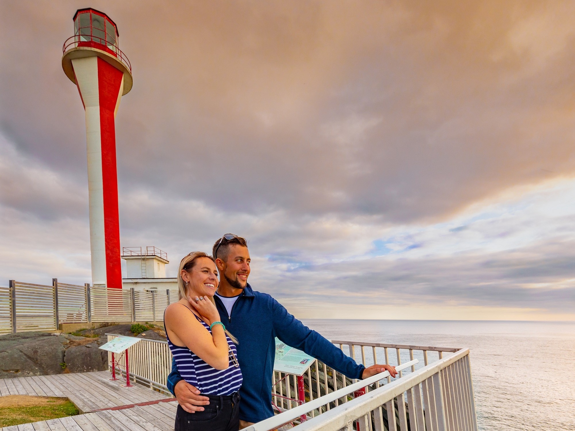 A couple looks out to the ocean with a red and white lighthouse in the background.