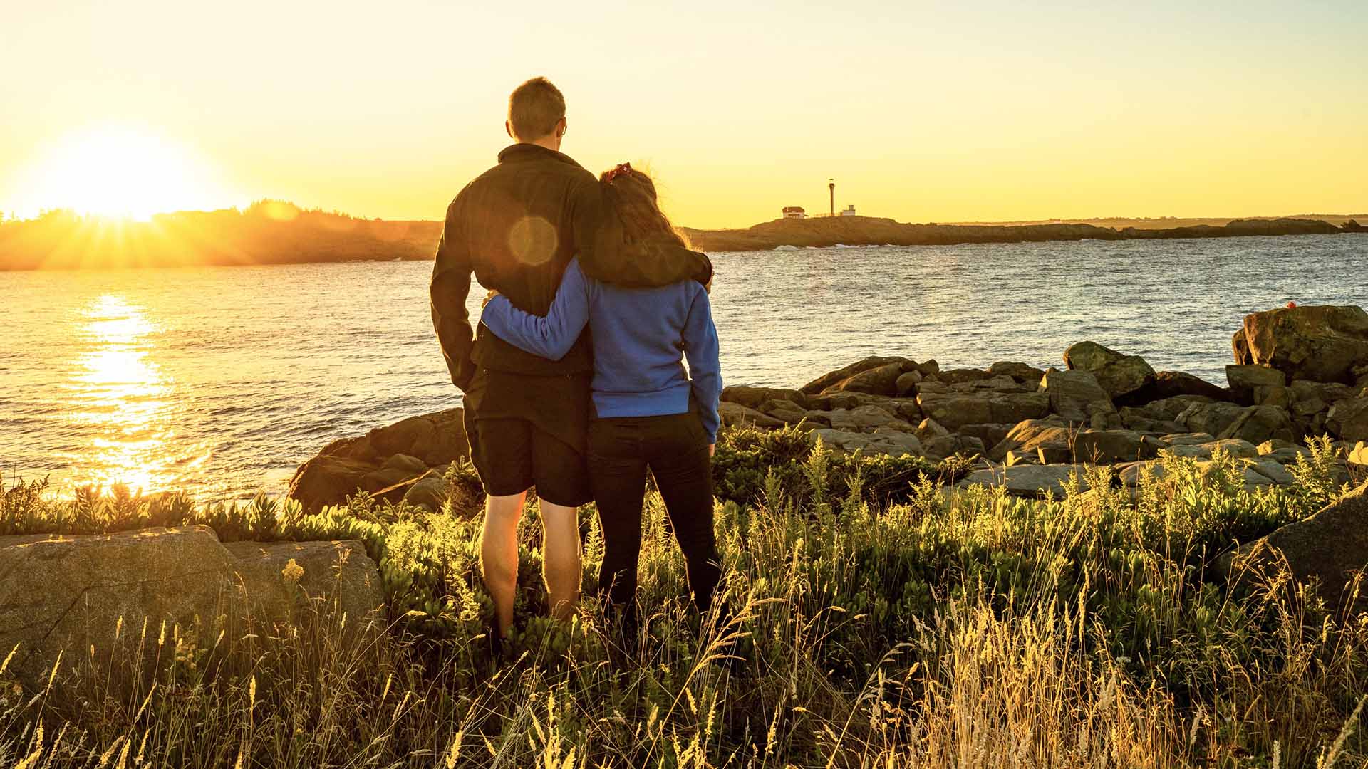 A couple stands by the ocean at sunset looking at the Cape Forchu Lighthouse