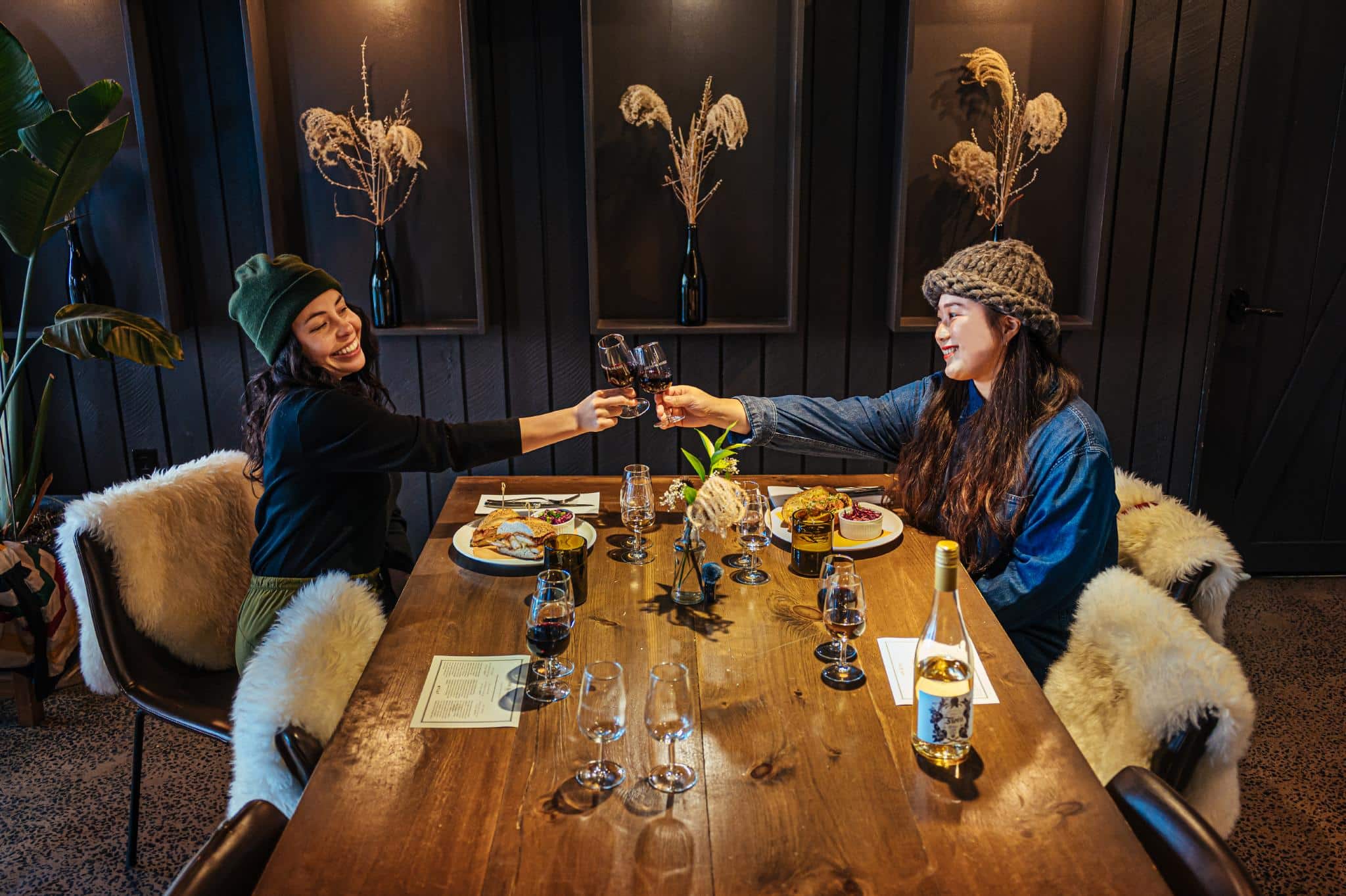 Two people cheers glasses of wine over a large wooden table. They both have meals in front of them.