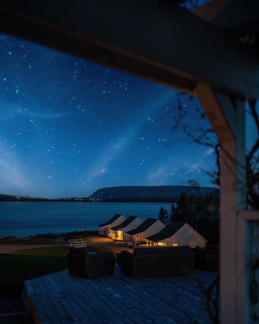 Three cottages by the water at night. There are lots of stars in the sky. The lights are on in the entrance to the cottages.