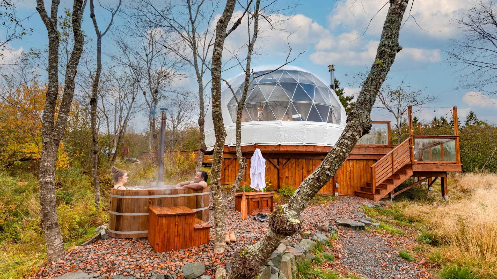 A white geodesic dome is perched above a couple in a hot tub beneath a blue sky on an autumn day.
