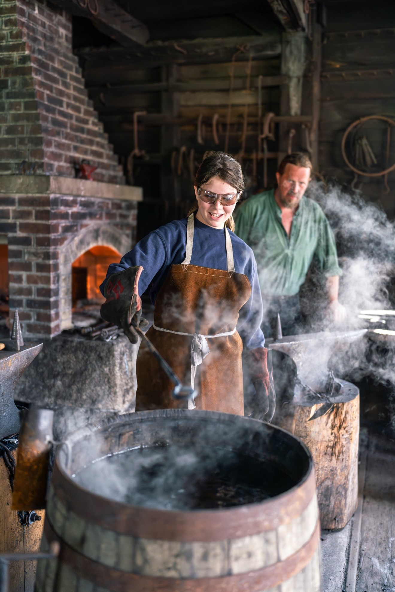 Two people take part in blacksmithing. One is wearing a heavy duty apron. The two people appear to be standing near a large brick fireplace.