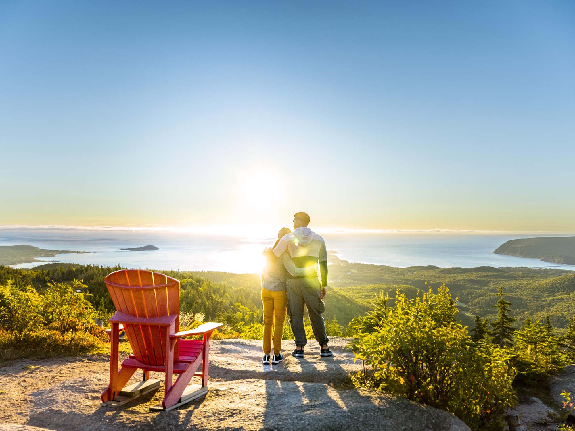 Two people embrace as they face the rising sun as they overlook the incredible views from the top of the Franey Trail in the Cape Breton Highlands National Park in Ingonish, Nova Scotia. A Parks Canada red chair is just behind them.