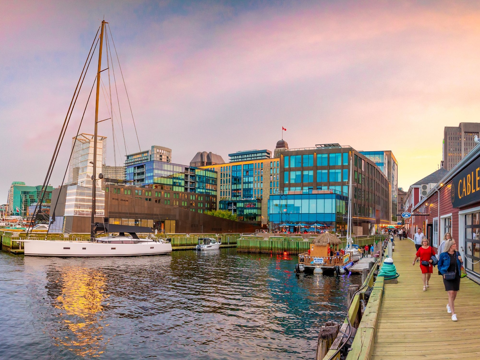 People walk along the boardwalk of a seaside city, with a sailboat docked nearby and city buildings in the background.