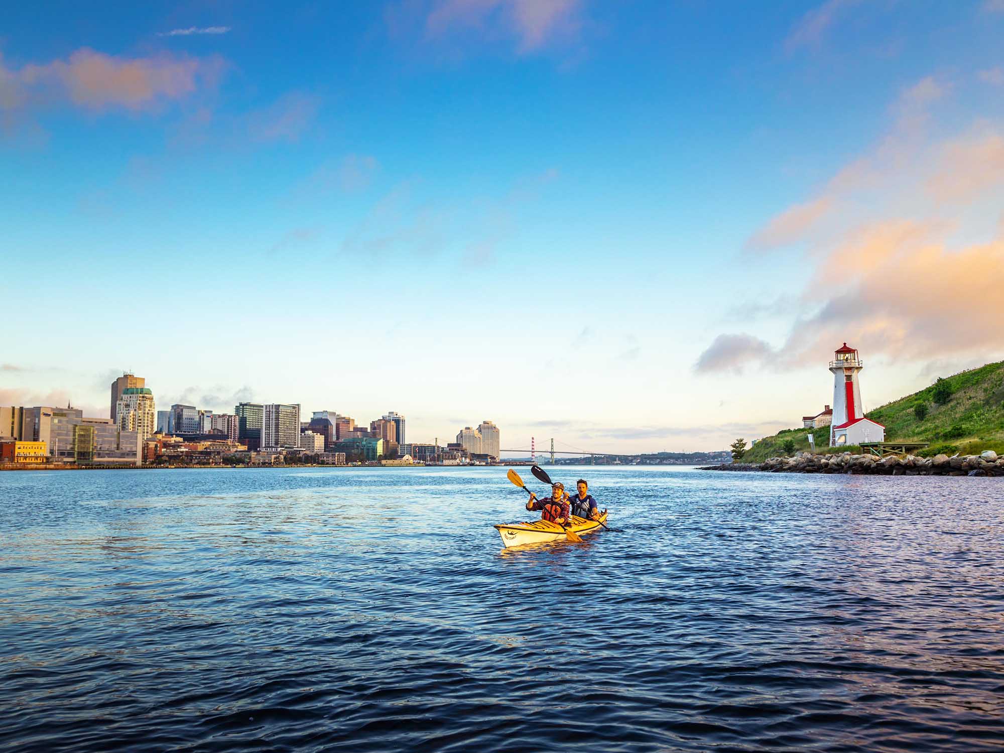 Two people in a tandem sea kayak p[addling next to Georges Island National Historic Site in Halifax Harbour with the skyline of downtown Halifax and the MacDonald Bridge behind them on a summer day in Nova Scotia.