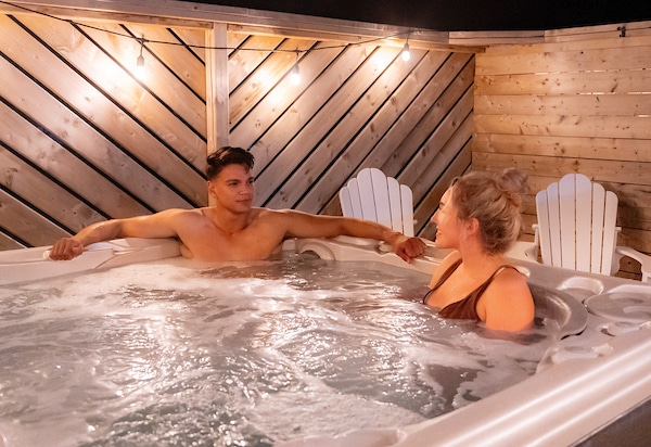 Two people sit in a hot tub. There appears to be a large wooden fence or wall surrounding them.