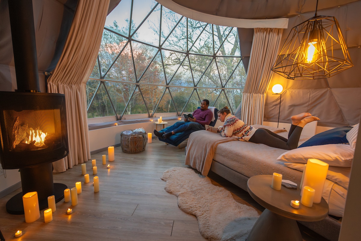 Two people relax, one in a chair and one on a bed, inside a dome. Out the window lots of trees can be seen. Inside the dome, dozens of flameless candles are placed around the floor and surfaces.