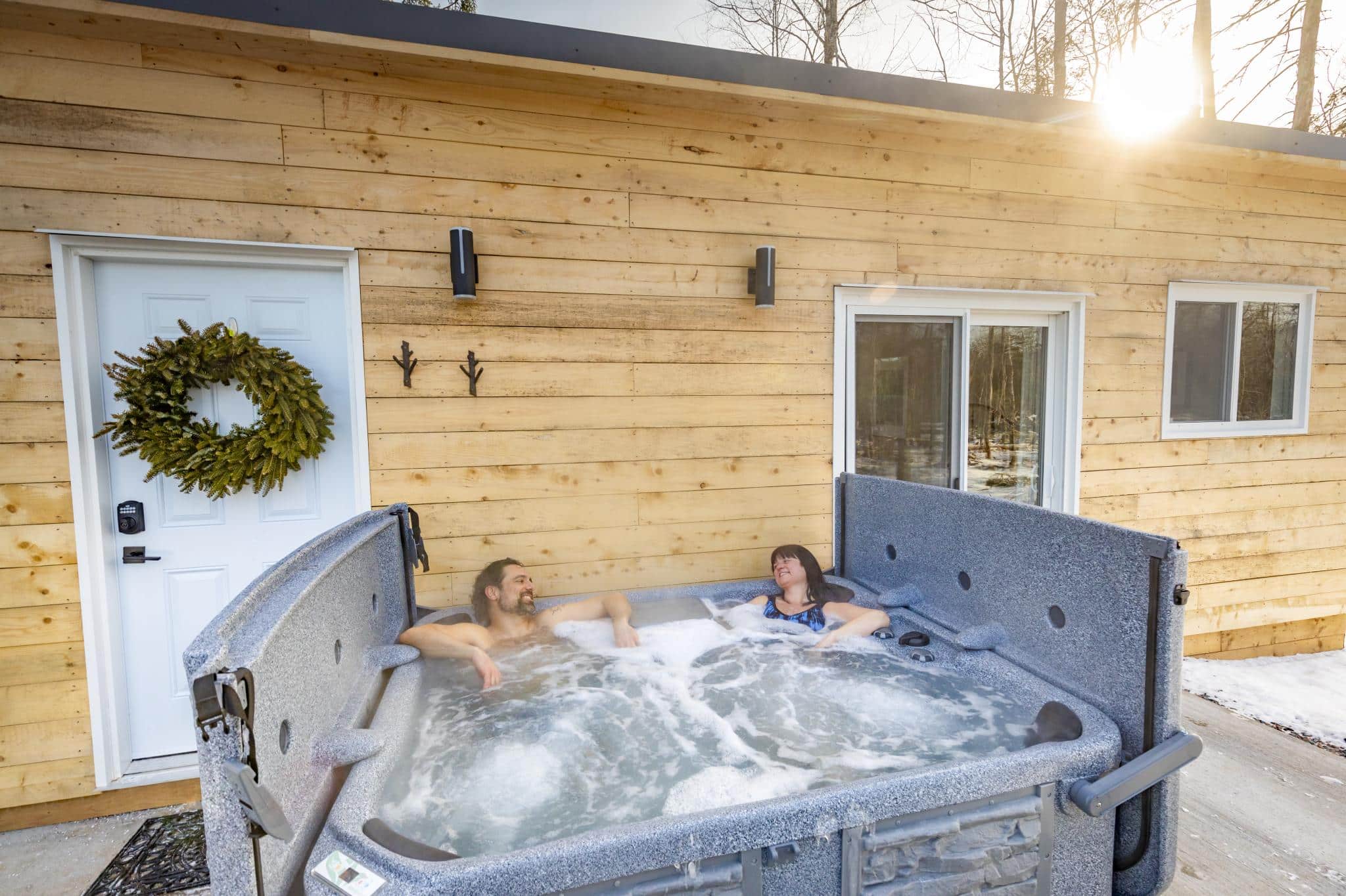 Two people sit in a hot tub at Seek Wilderness Resort. There are sides to the hot tub for privacy. Directly behind them in a cabin with wooden siding.