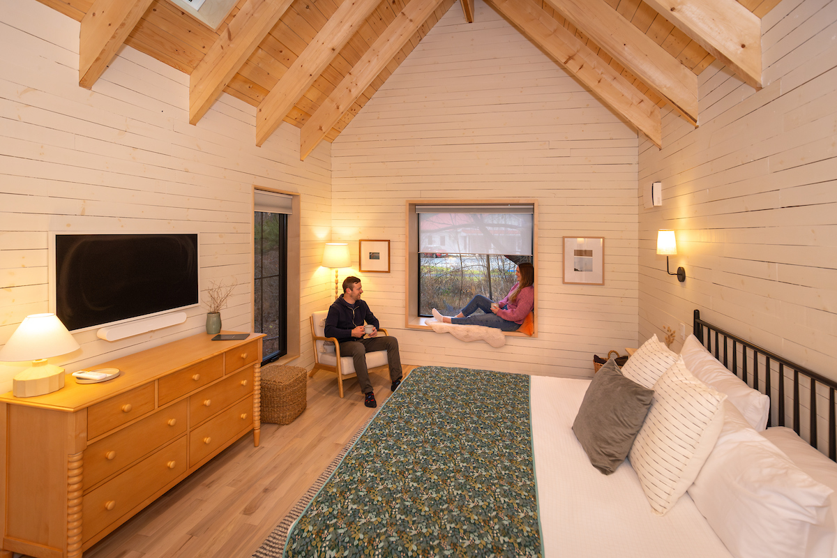 The interior of a cabin can be seen. Two people sit by a window, with one sitting in a windowsill. The ceiling is an A-Frame. There is a wooden dresser and large bed.