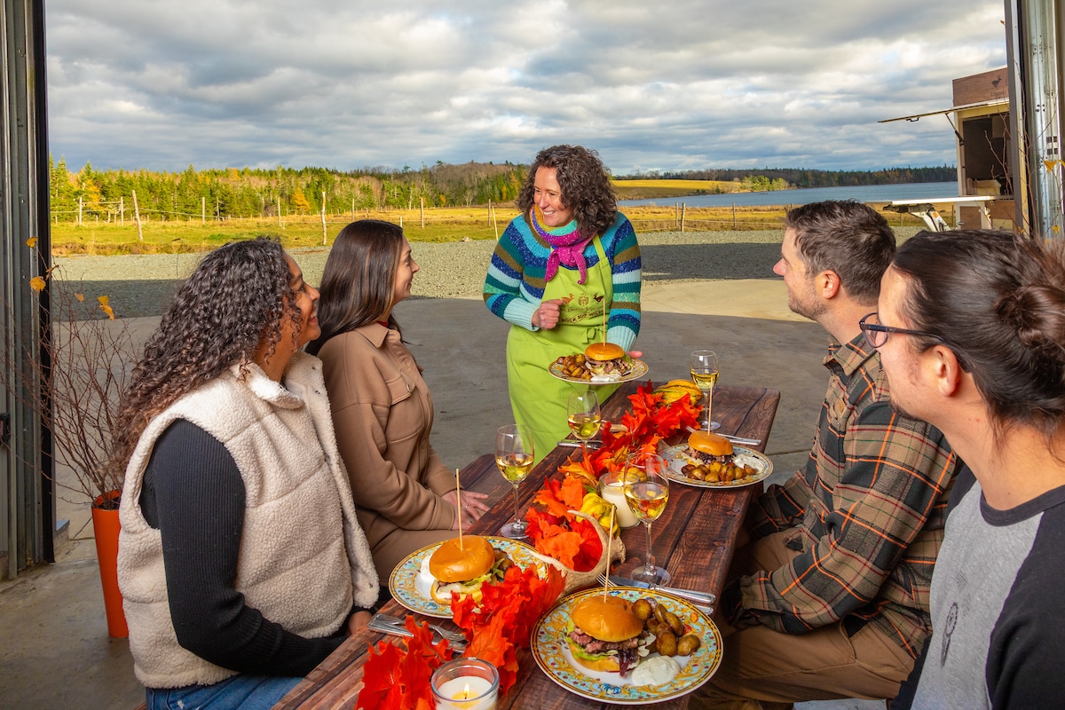 A group of four people sit at a table outside. A fifth person is serving their meal - burgers and home fries. In the background there are trees and a lake.