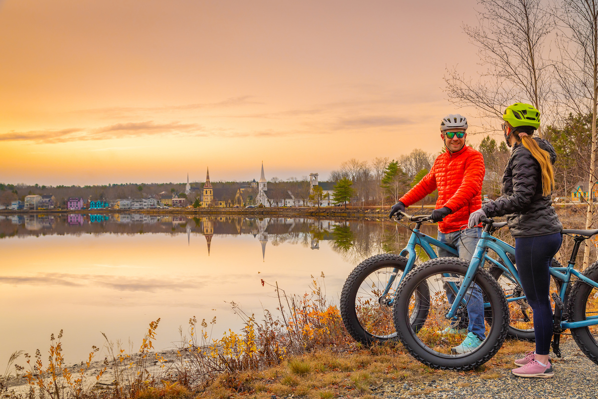 Two people pose with fat bikes on the edge of a body of water. In the background is the town of Mahone Bay, with the iconic image of the three churches dotted along the waterfront.