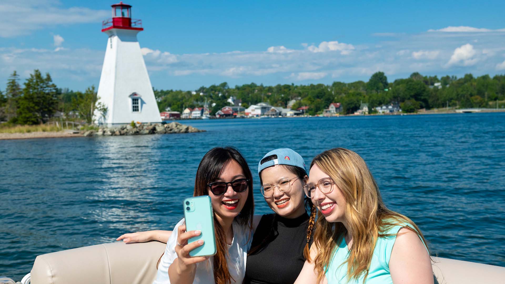 Friends talking a selfie of themselves with the Kidston Island lighthouse and the town of Baddeck behind them in Nova Scotia