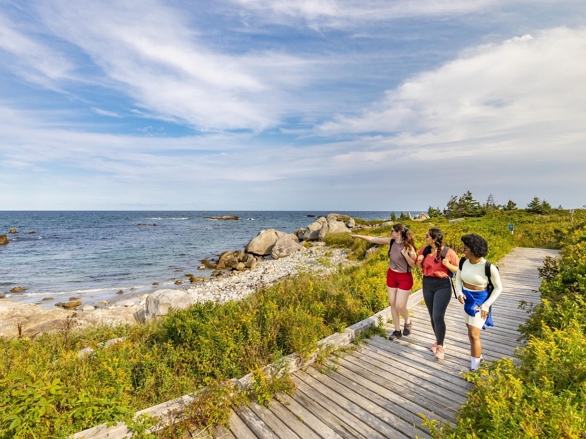 Three women hike along a wooden boardwalk next to the ocean.