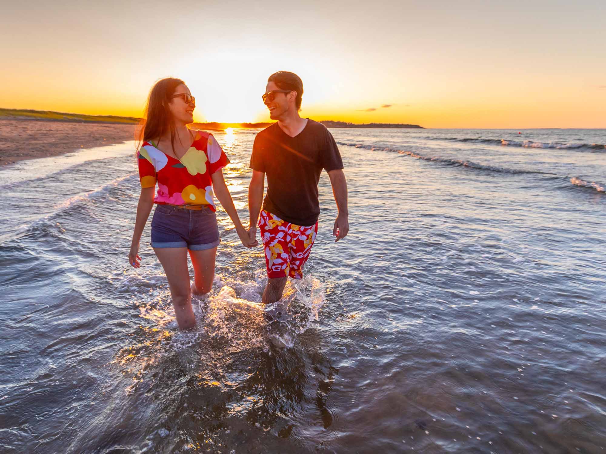 Two people holding hands and laughing as they look at each other as they walk in the warm ocean surf at sunset at Melmerby Beach Provincial Park near New Glasgow Nova Scotia.