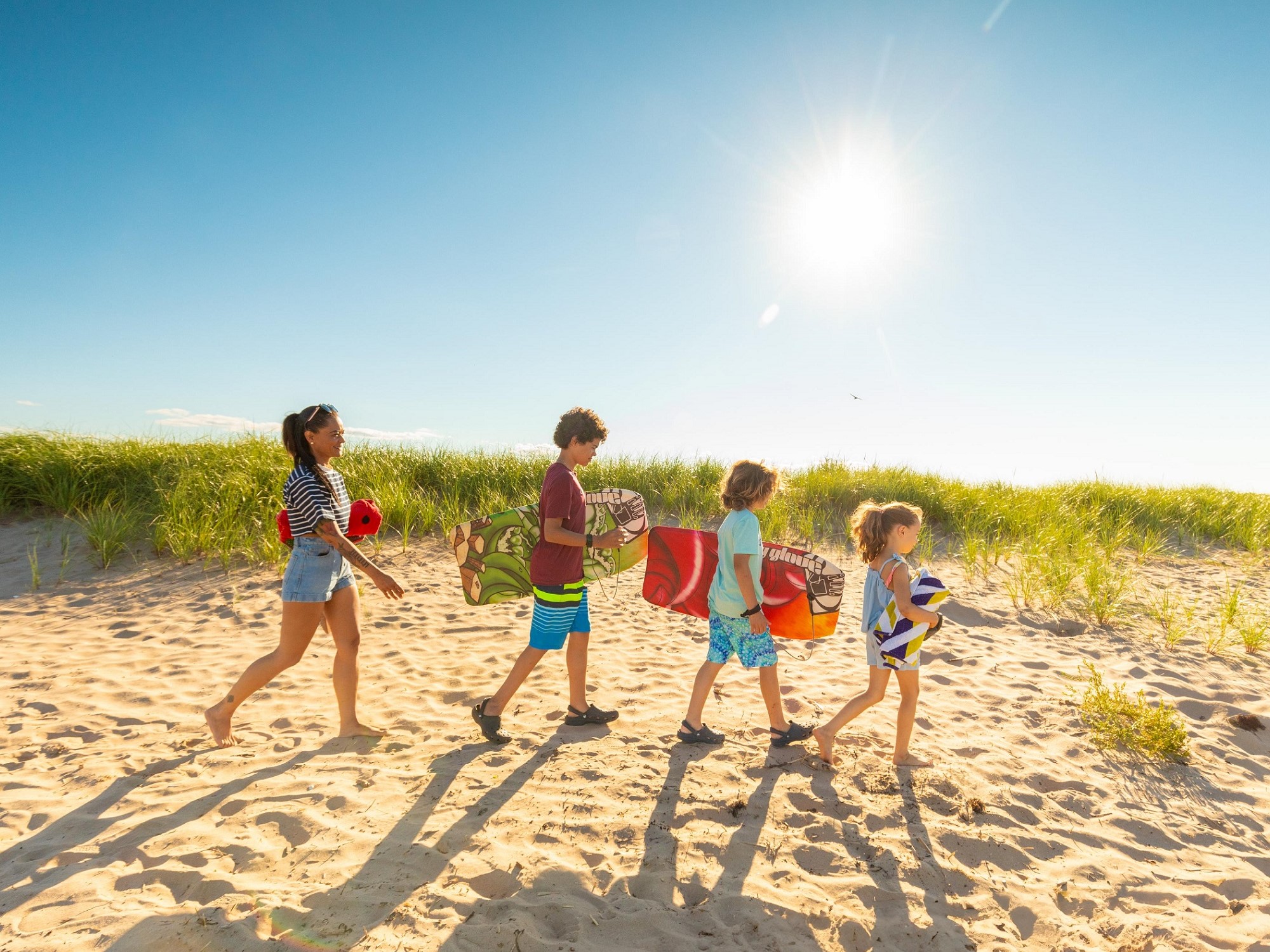 A family walks along the sand on a sunny day carrying their gear for a beach day.