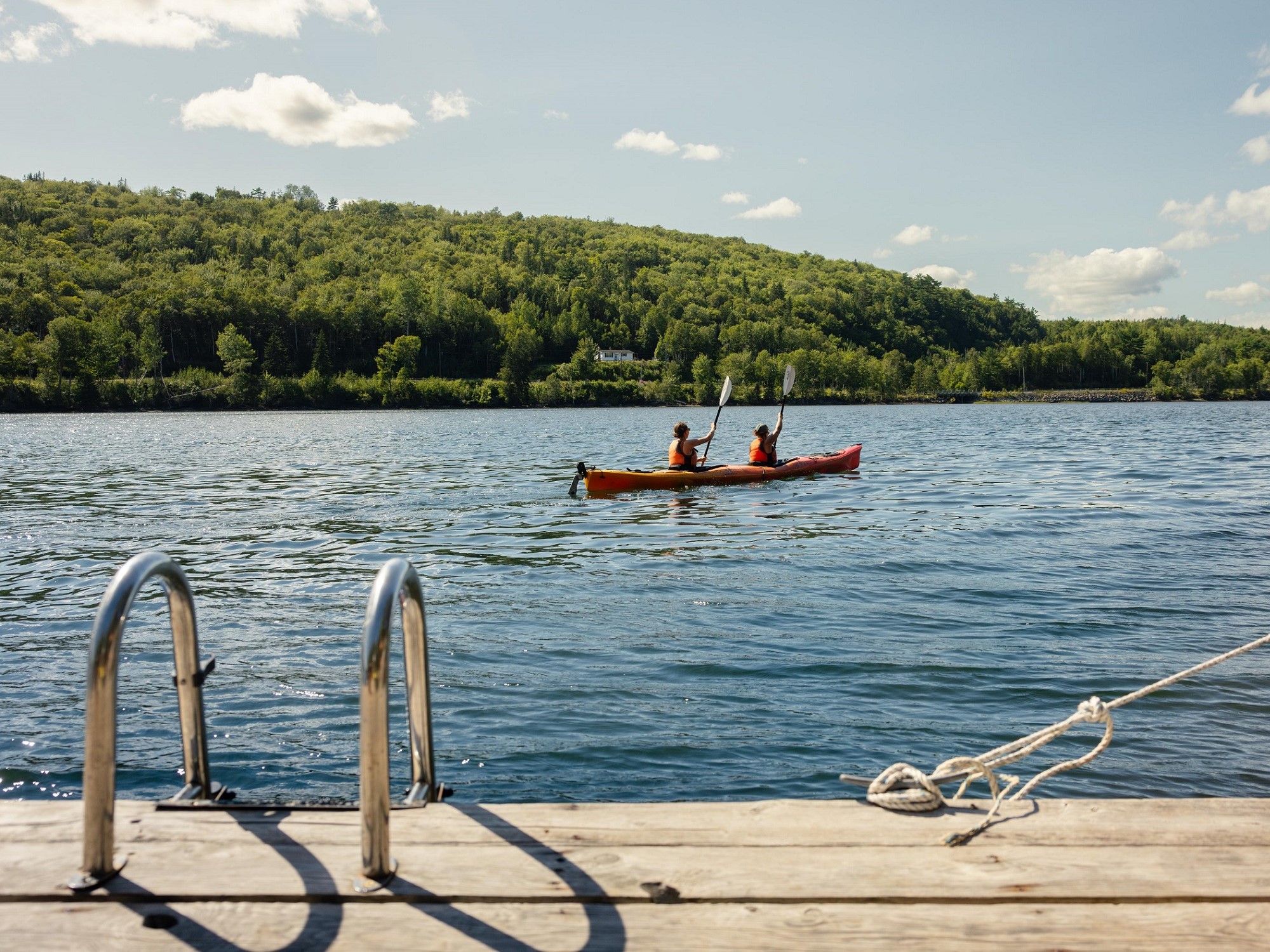 Two people paddle in a double kayak with green highlands in the background on a blue sky day.