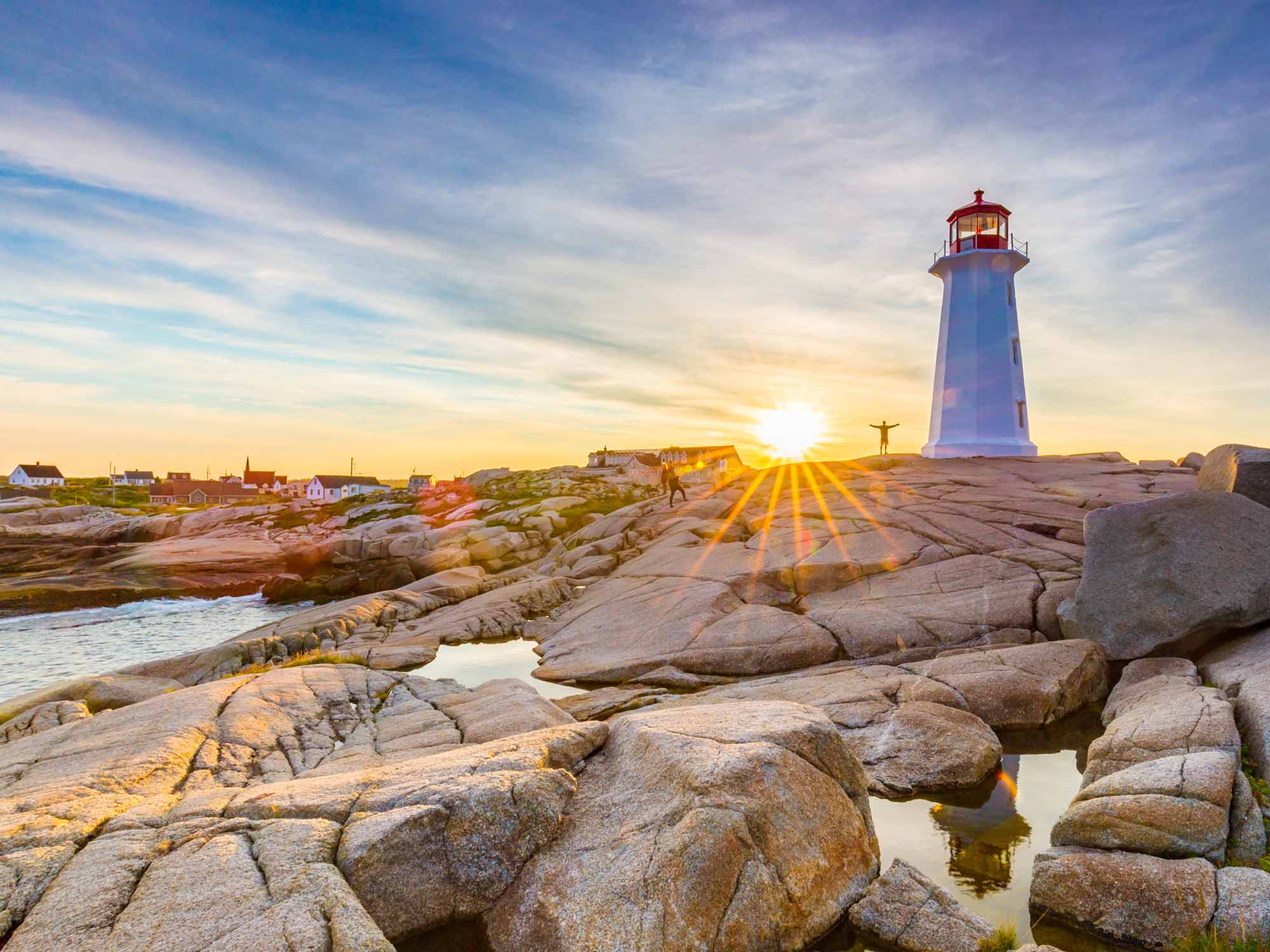 A person standing with their arms outstretched to their sides as they stand next to the Peggy's Cove Lighthouse at sunrise in Peggy's Cove in Nova Scotia