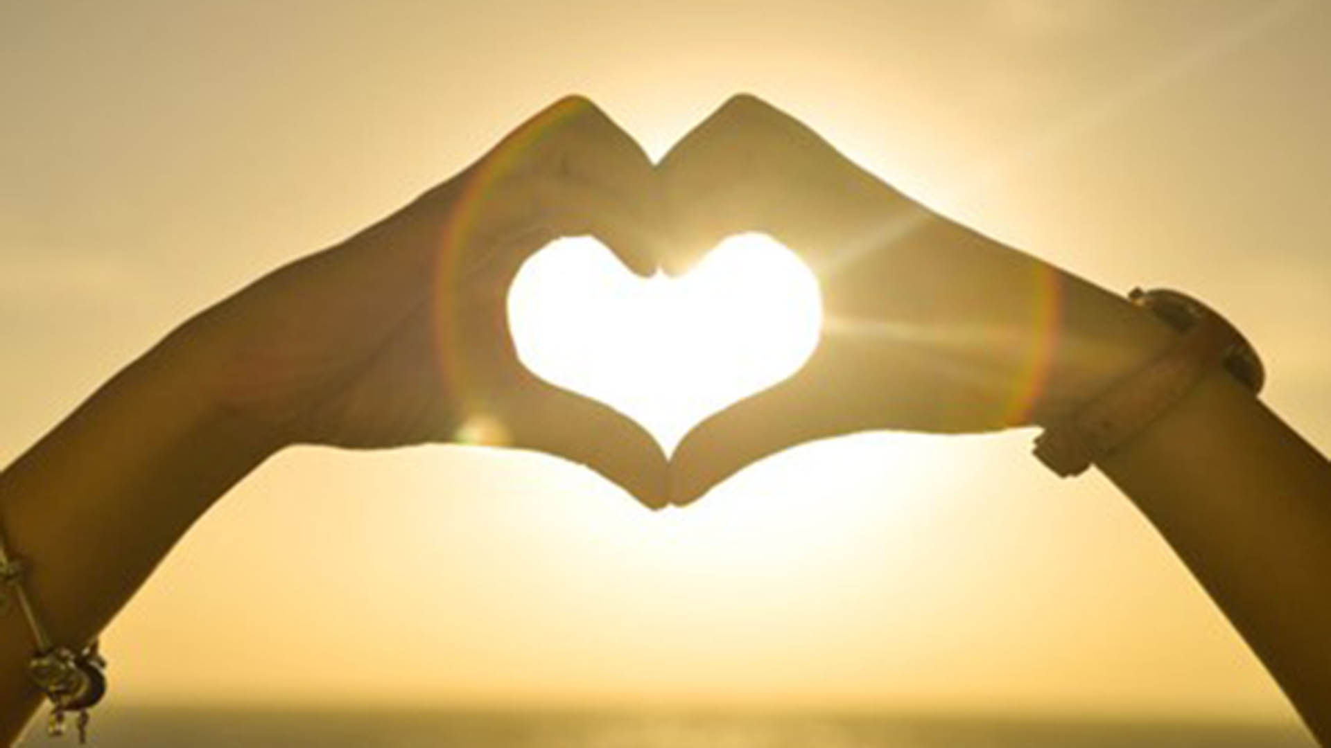 Two hands join together to create the shape of a heart during sunset with the light shining through their hands