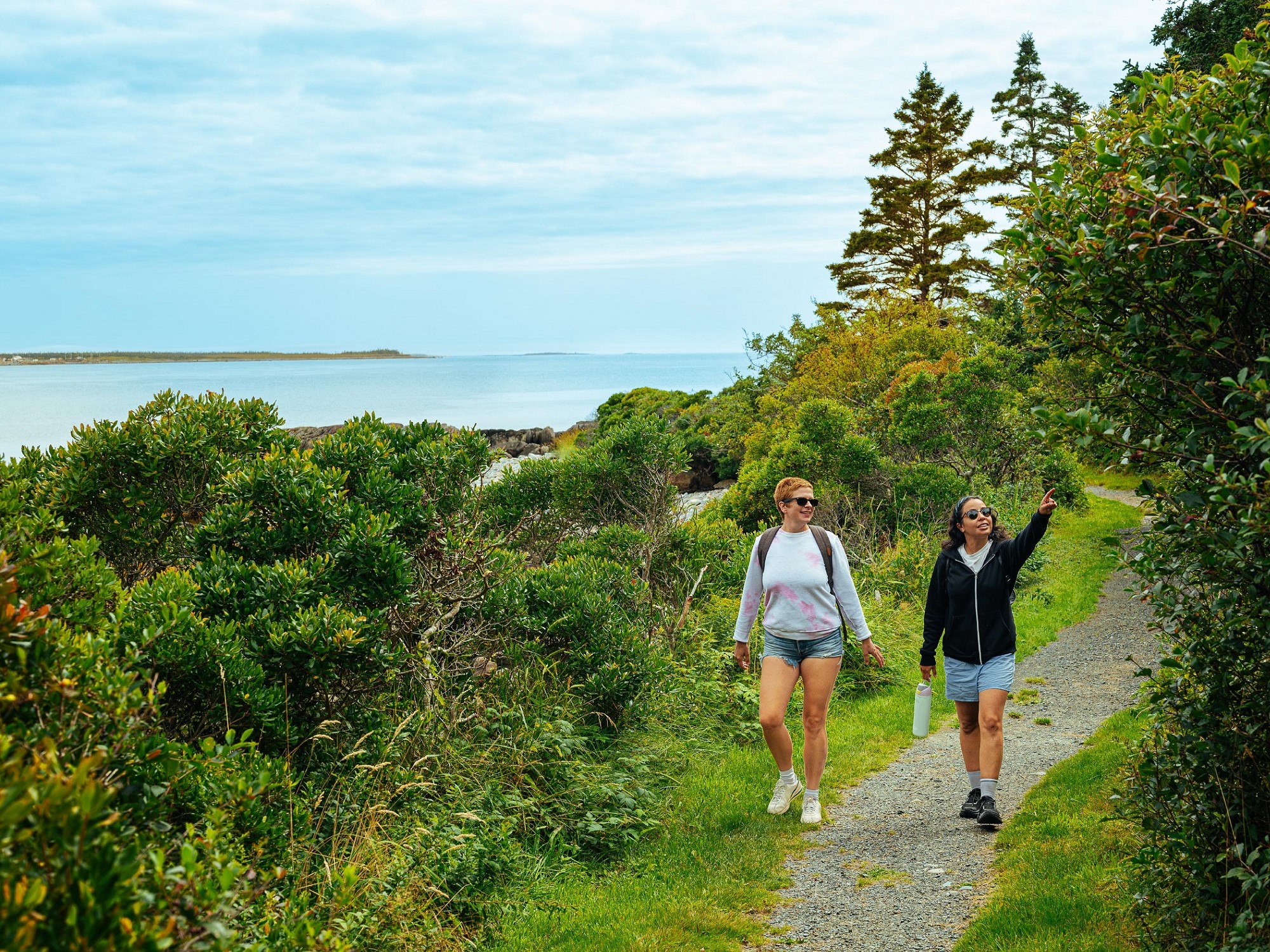 Two women walk along a waterside trail, one is pointing to a tree.