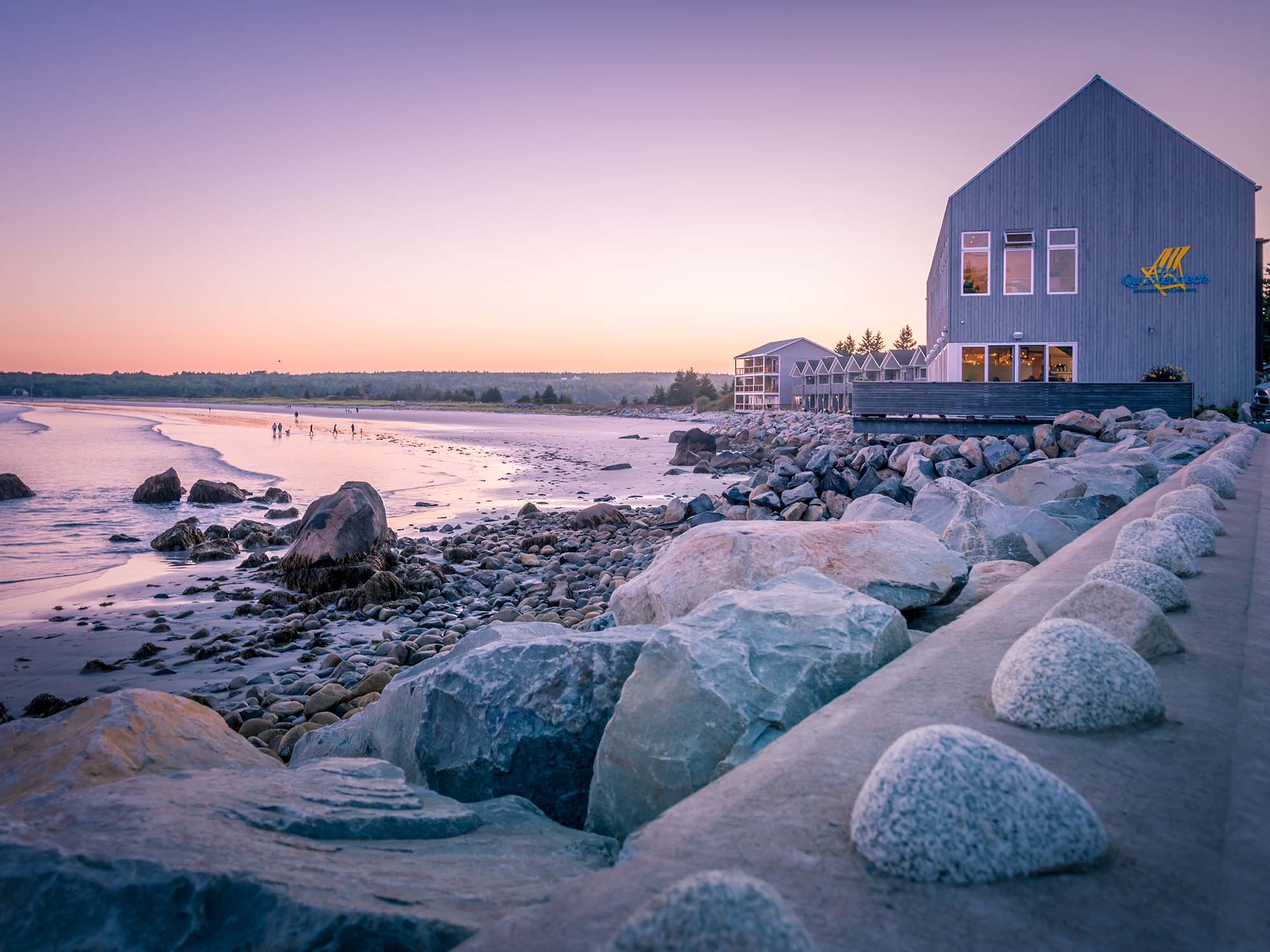 View of The Quarterdeck Beachside Villas and Grill at sunset on a calm summer evening with a purplish hue from the setting sun over the beach and ocean and buildings in Nova Scotia