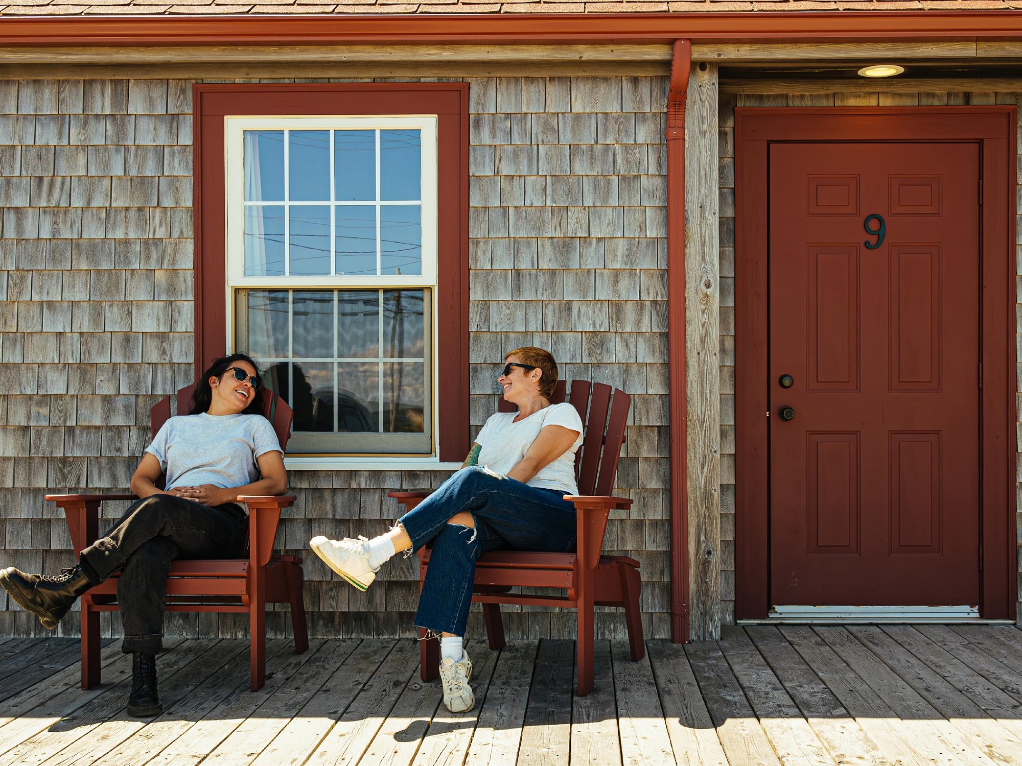 Two women sit in red chairs in front of a red motel door of a cedar shake building.