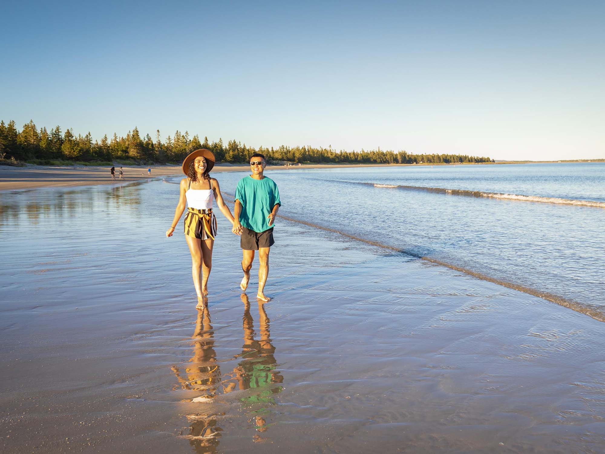 Two people holding hands and smiling as they walk in the calm surf at Rissers Beach Provincial Park on a warm summer day in Nova Scotia.
