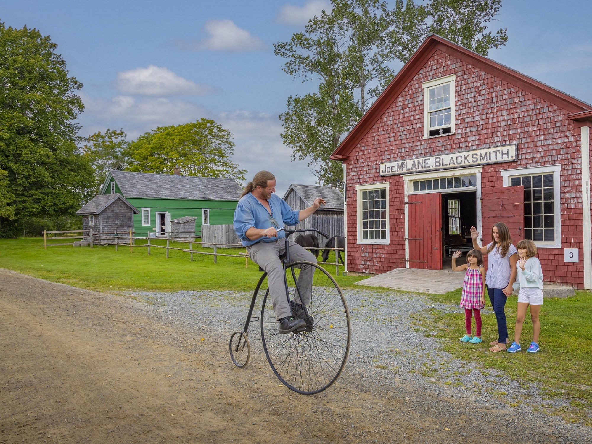A mom and two kids wave at a man riding an old fashioned bicycle at Sherbrooke Village Museum.