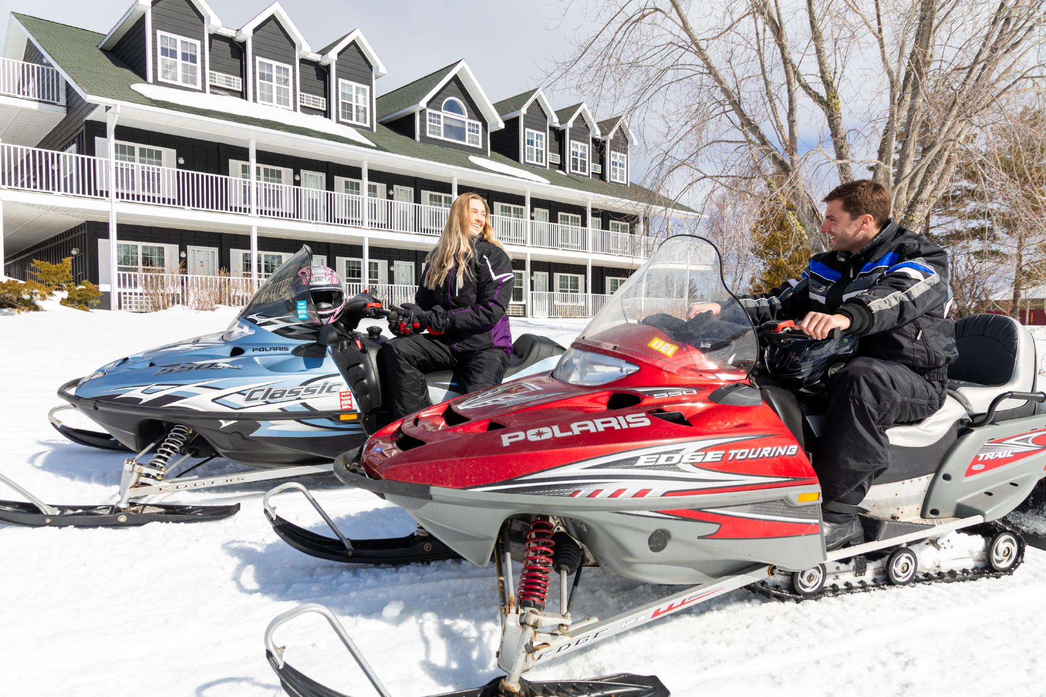 Two people sit on snowmobiles in front of Inverary Resort.