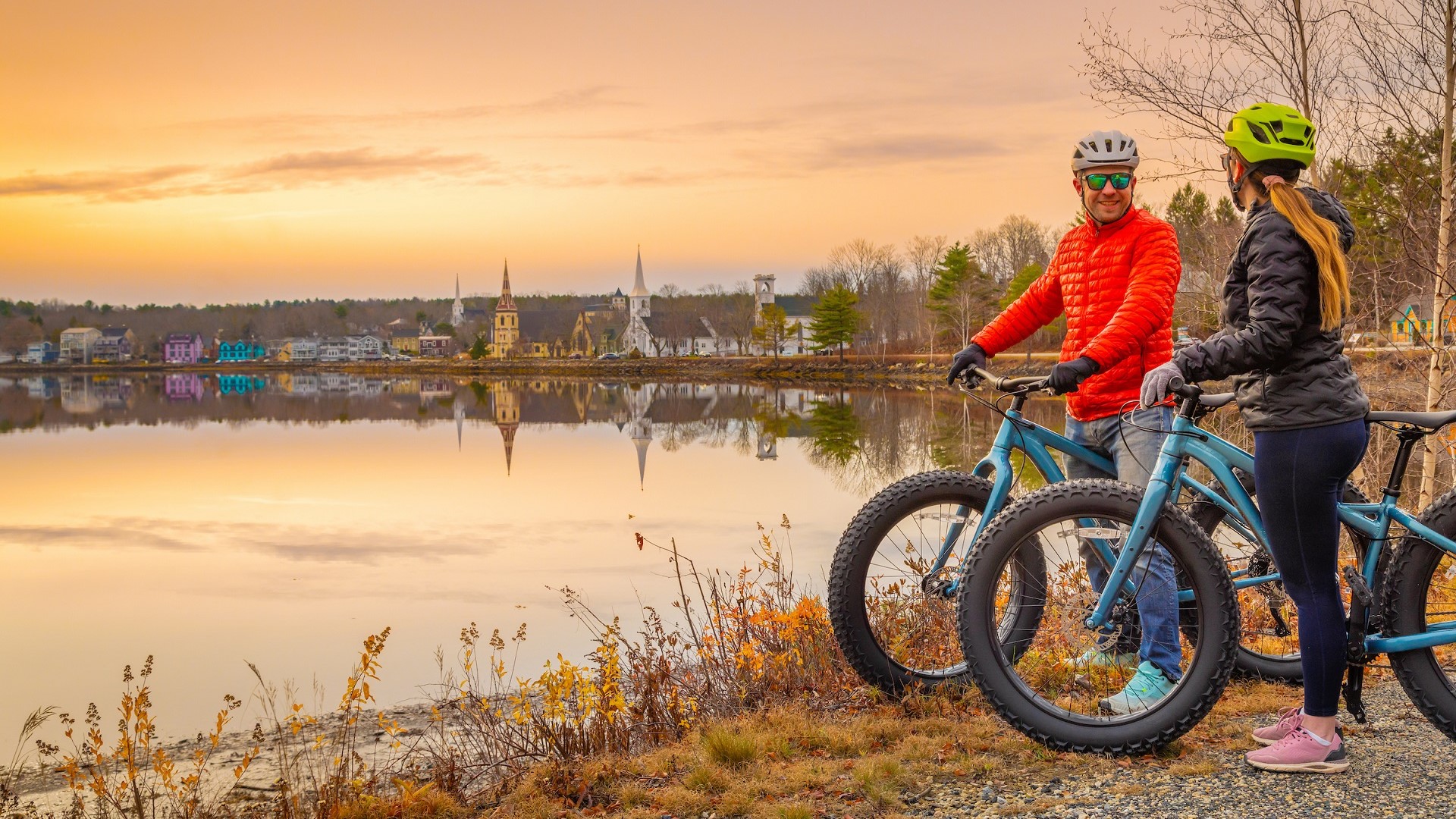Two people on bikes take a rest along the water's edge overlooking the iconic Three Churches in Mahone Bay during an autumn day