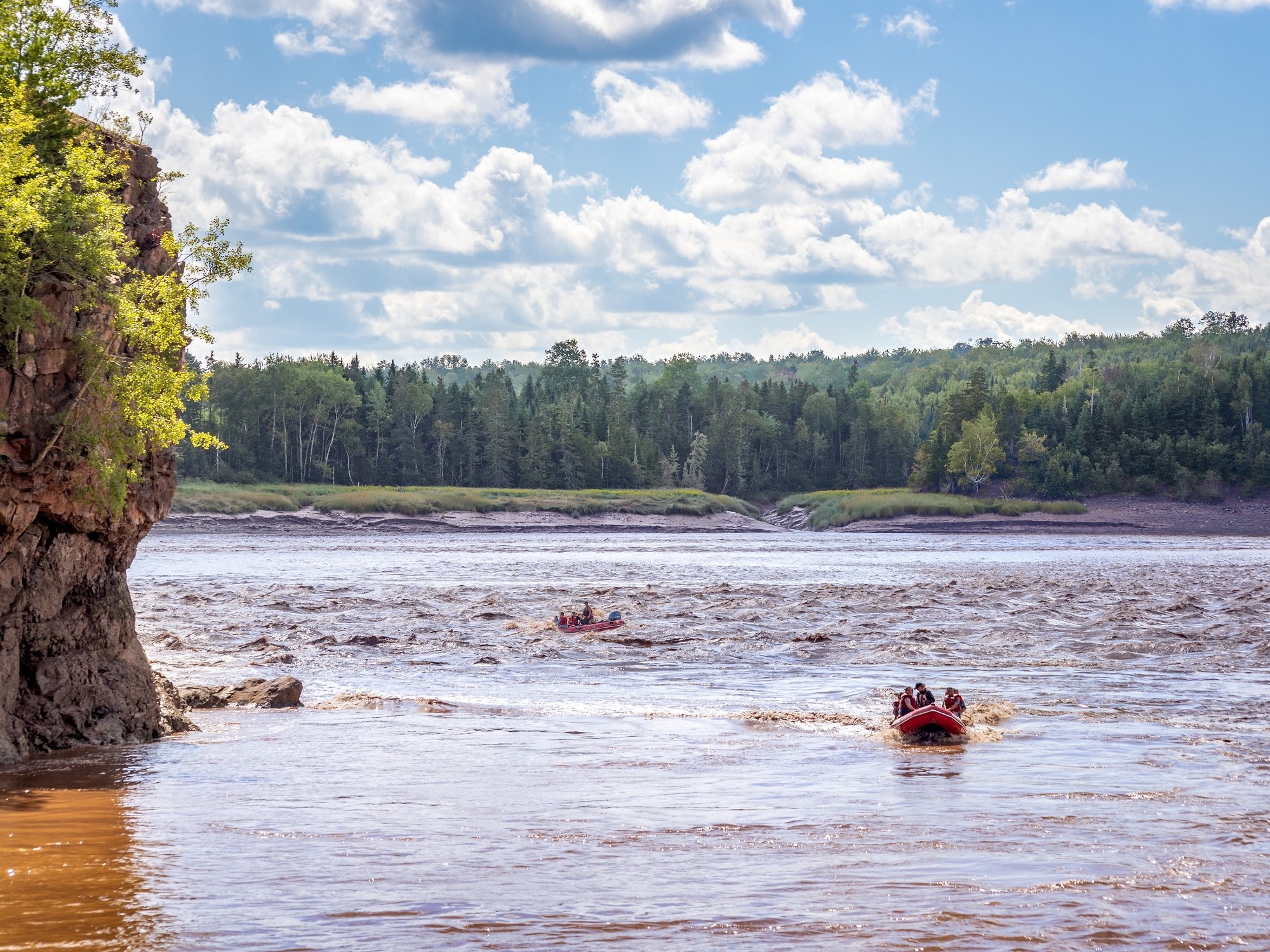 A tidal bore rafting boat sails down Shubenacadie River alongside cliffs rising from the water