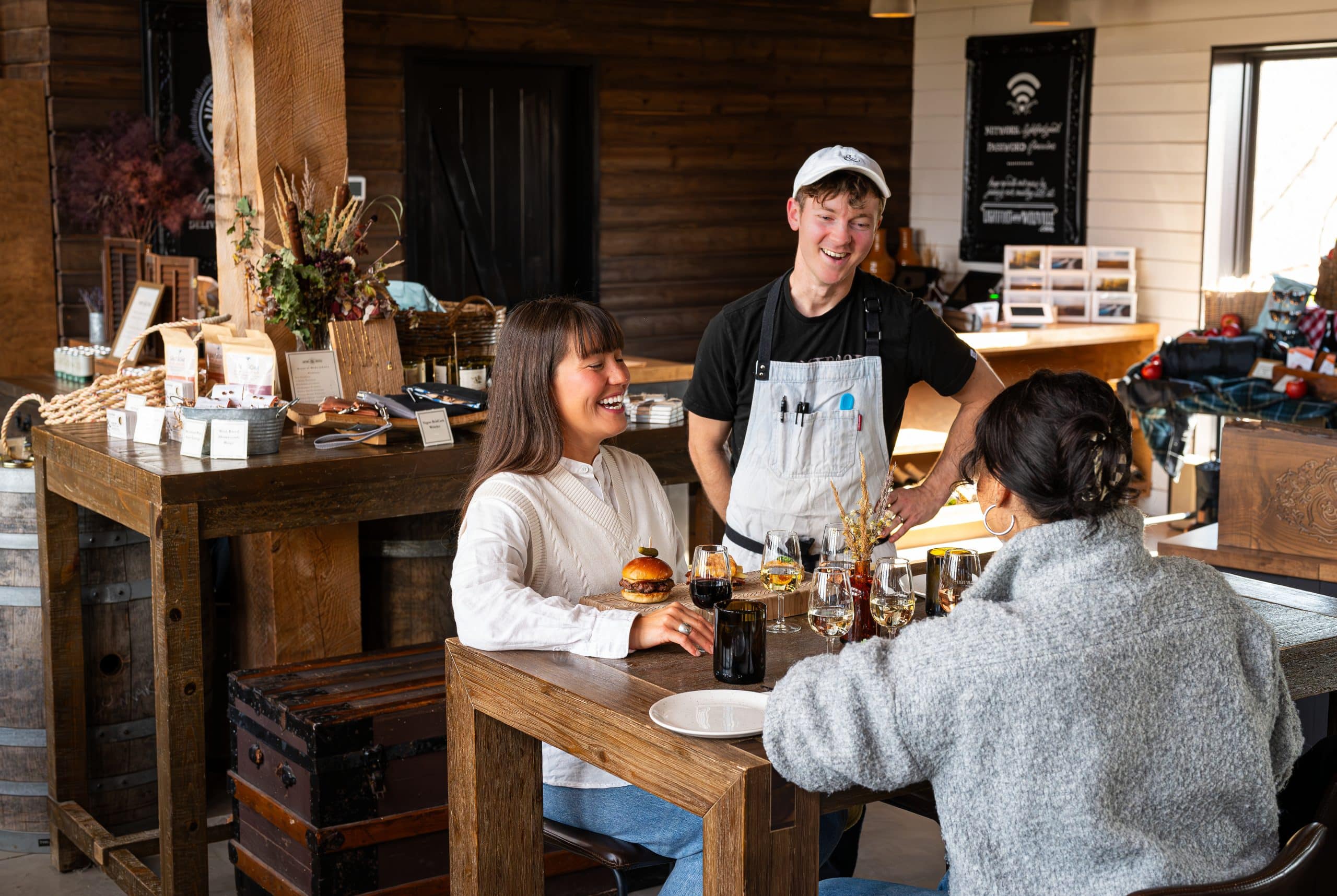 A chef at a winery talks to two people dining. The patrons both have wine glasses and are eating.
