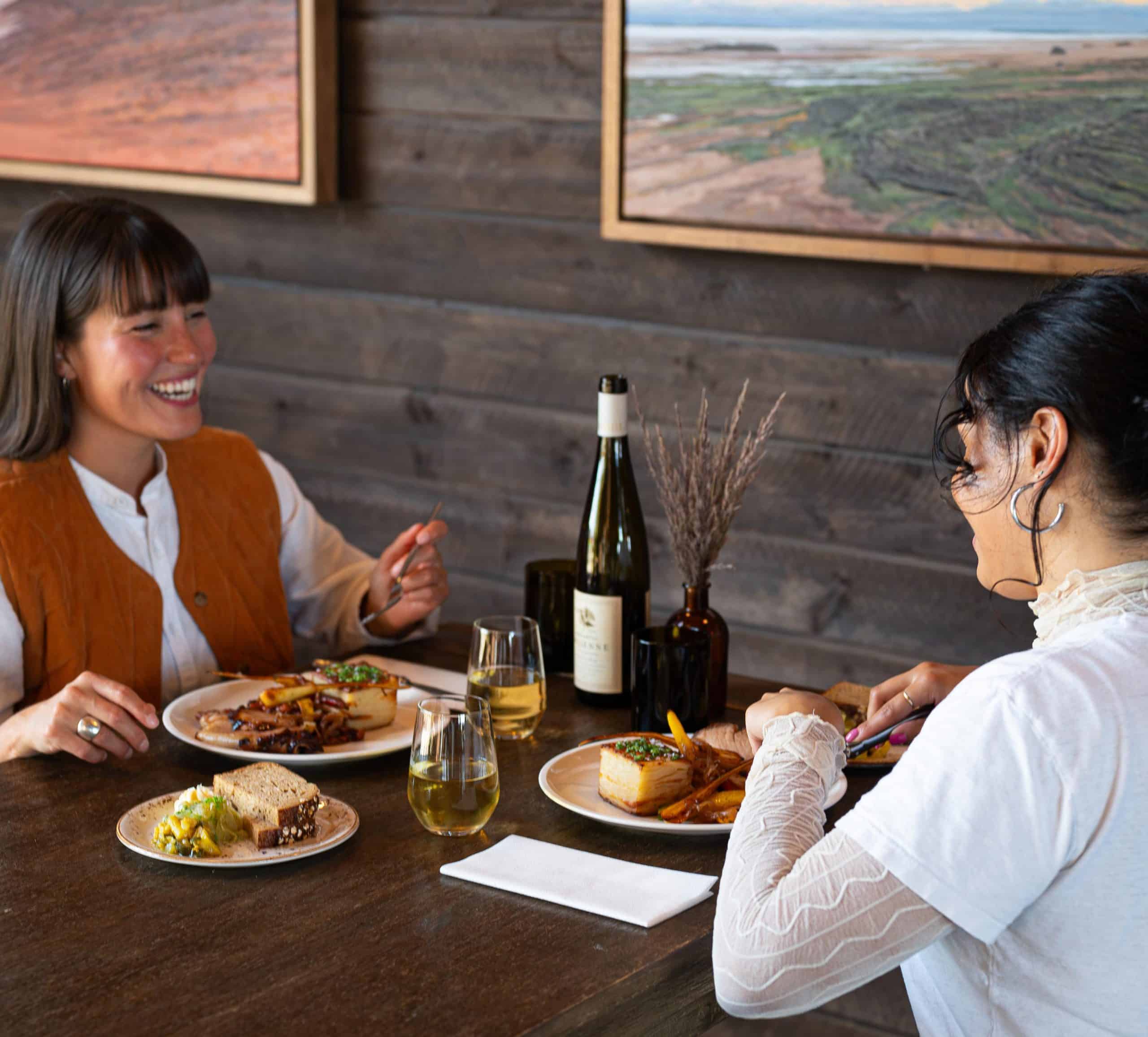 Two people sit at a wooden table with art in the background. They are enjoying a ham dinner with a wine pairing and smiling at one another.