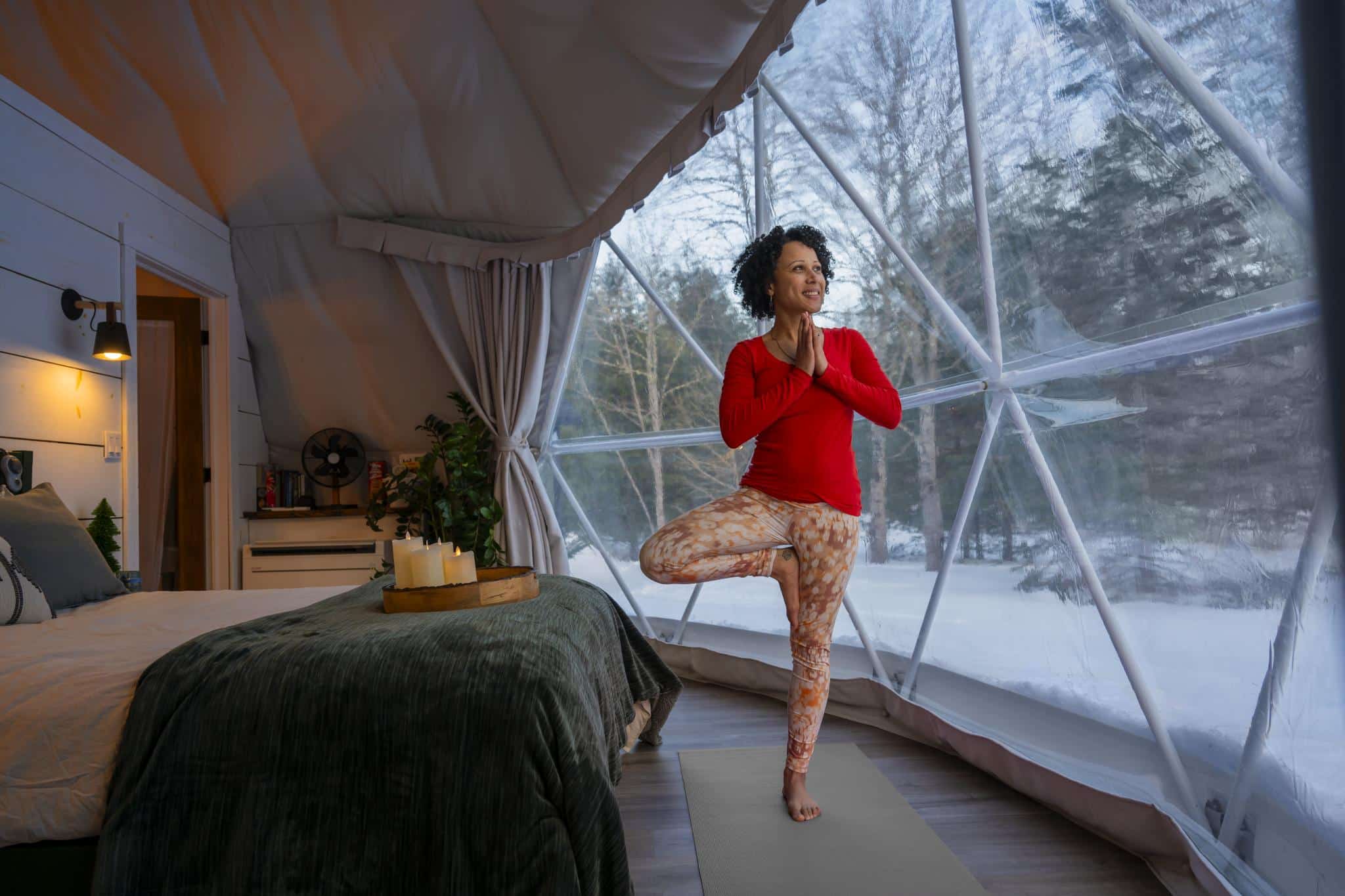 A person does yoga inside a dome. You can see large windows behind the person and outside it is winter and there is lots of snow.