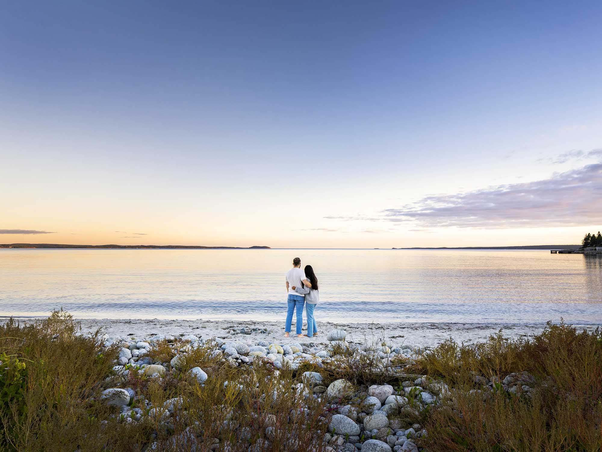 Two people embrace standing barefoot facing the water on the beach at Wilson's Coastal Club Resort in Boutiliers Point, Nova Scotia just as the sun has set on a summer's day.