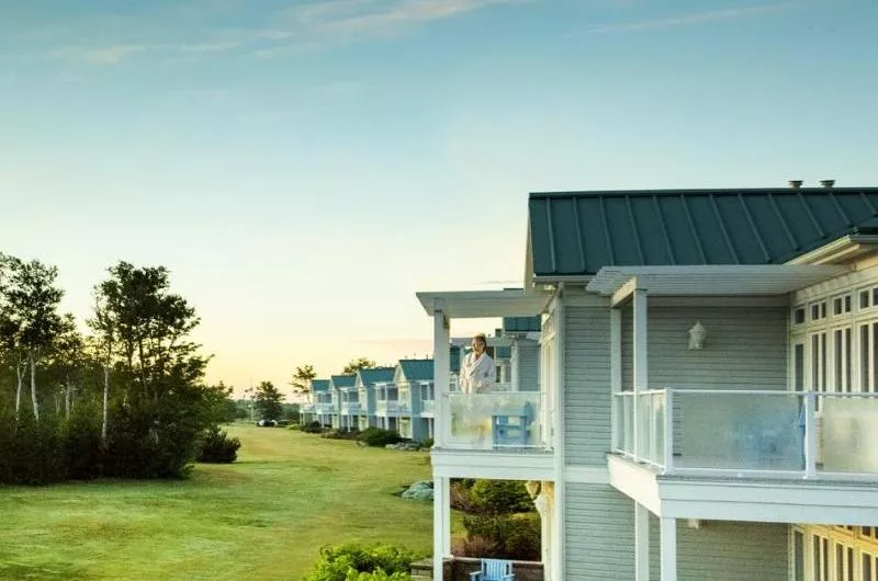 A person in a white housecoat stands on a balcony of a house that is one of many in a row.