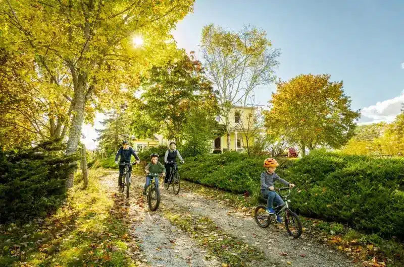 A family of four, two parents and two children, bikes along a trail. It is a sunny day and the trees and grass are lush. There is a yellow house in the background.