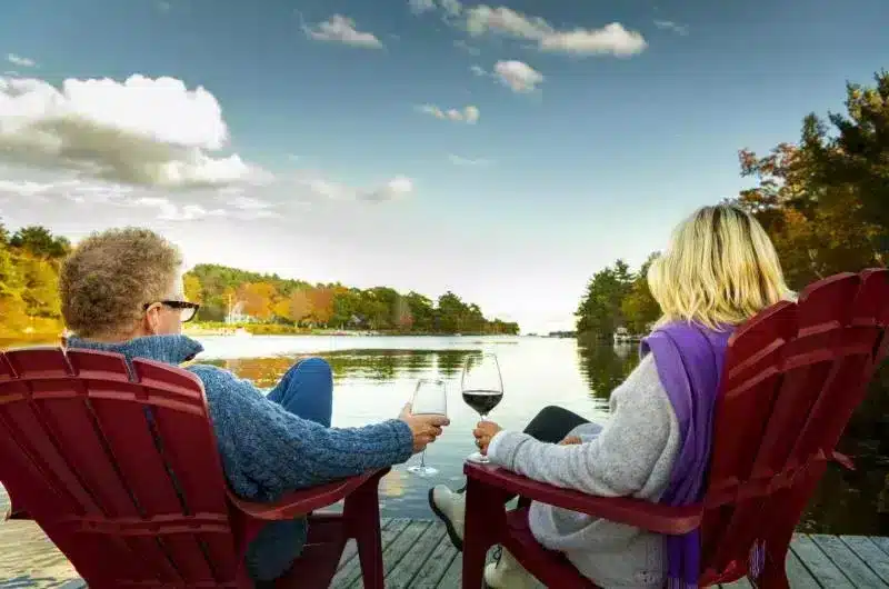 Two people sit on a dock in red Adirondack chairs overlooking the calm water. Both people hold glasses of wine. It is a sunny day with a blue sky and few clouds.