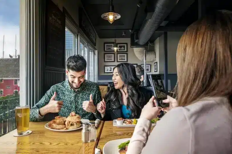 Three people are sitting in a booth at a restaurant. One person is taking a photo of the other two people. They have ordered their food and are excited to eat.